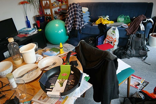 A living room cluttered with dirty dishes, grocery bags, back packs, exercise equipment, and clothing.